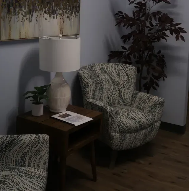 Cozy waiting area with patterned armchair, side table, lamp, and plant. A framed artwork hangs above.