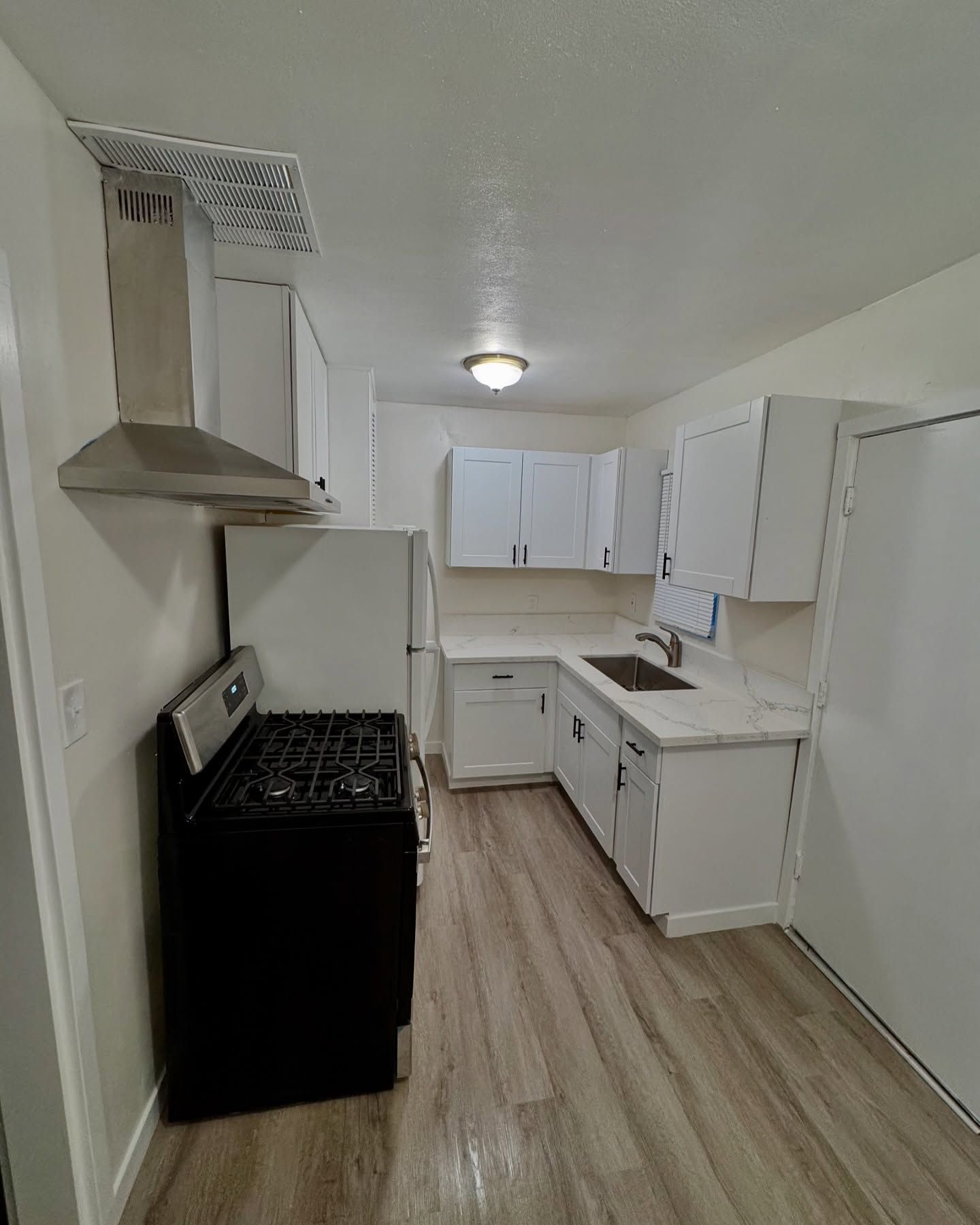 Small, white kitchen with appliances, cabinets, and a sink. Black stove and stainless steel hood.