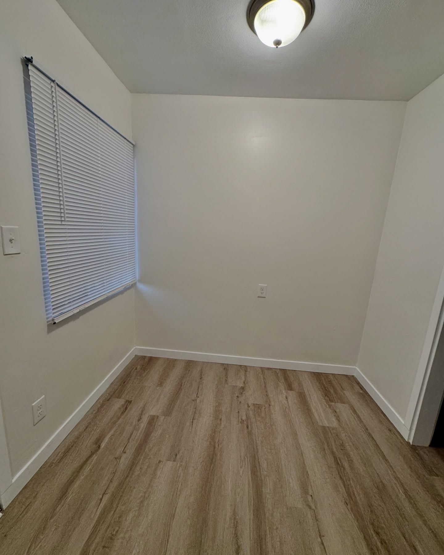 Empty room with beige walls, wood-look flooring, window with blinds, and a ceiling light.