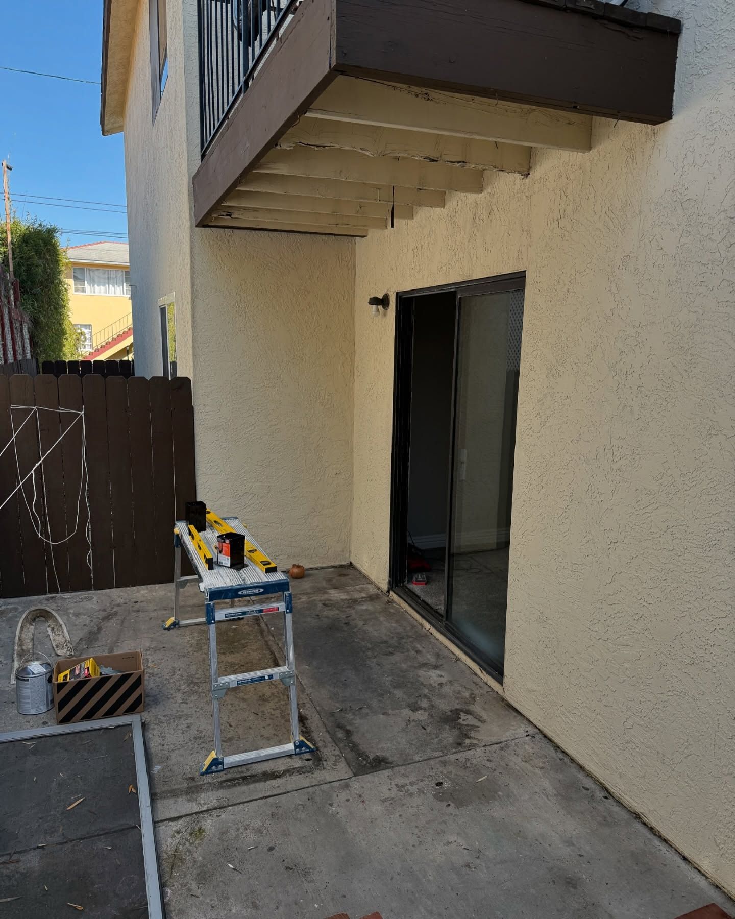 Back patio with tools, sliding glass door, and brown balcony. Brown fence on the left and stucco building in the background.