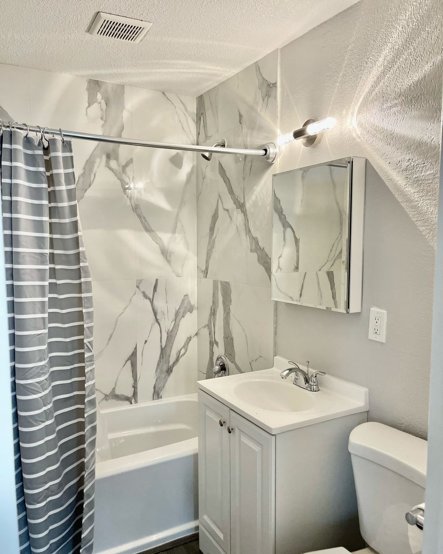 Small bathroom with white and gray marble-look shower tile, white sink and toilet, gray striped shower curtain.