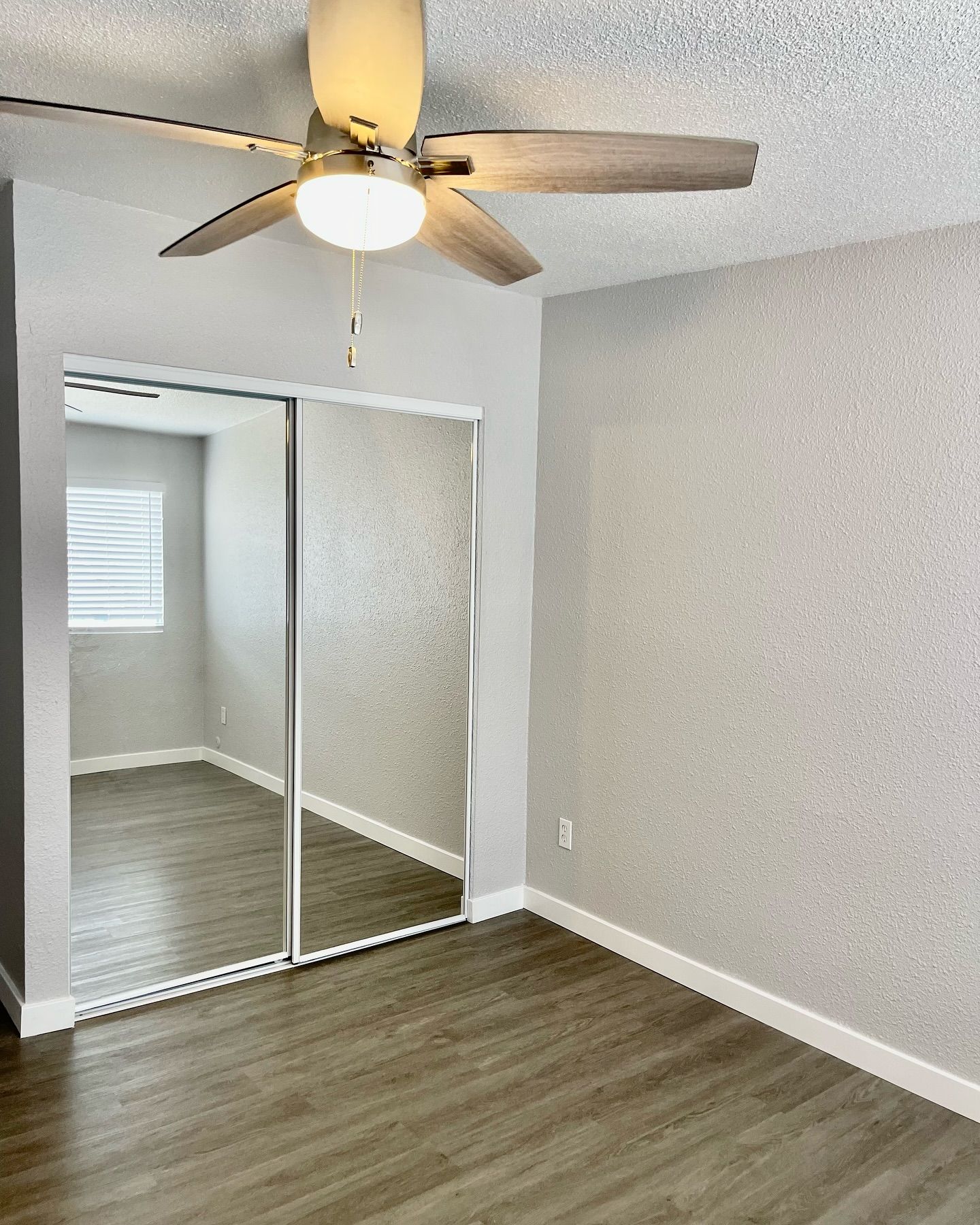 Empty bedroom with mirrored closet doors, ceiling fan, and gray walls, dark wood-look floor.