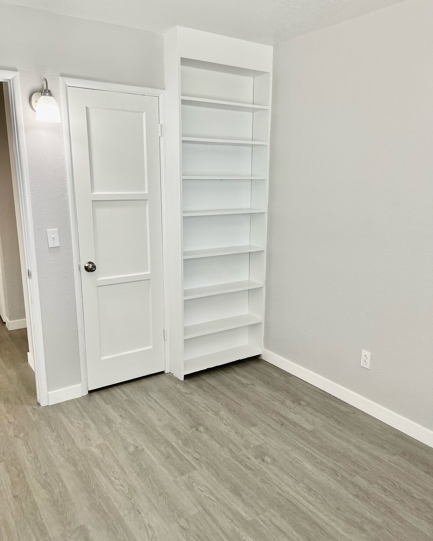 A room with a white door, a tall white bookshelf, and gray flooring and walls.