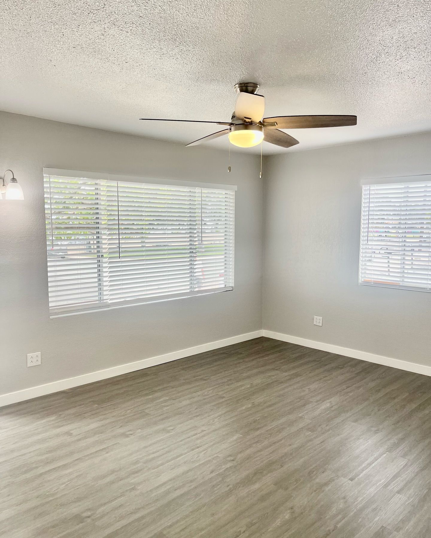 Empty room with gray walls, light wood-look floor, windows with blinds, ceiling fan.