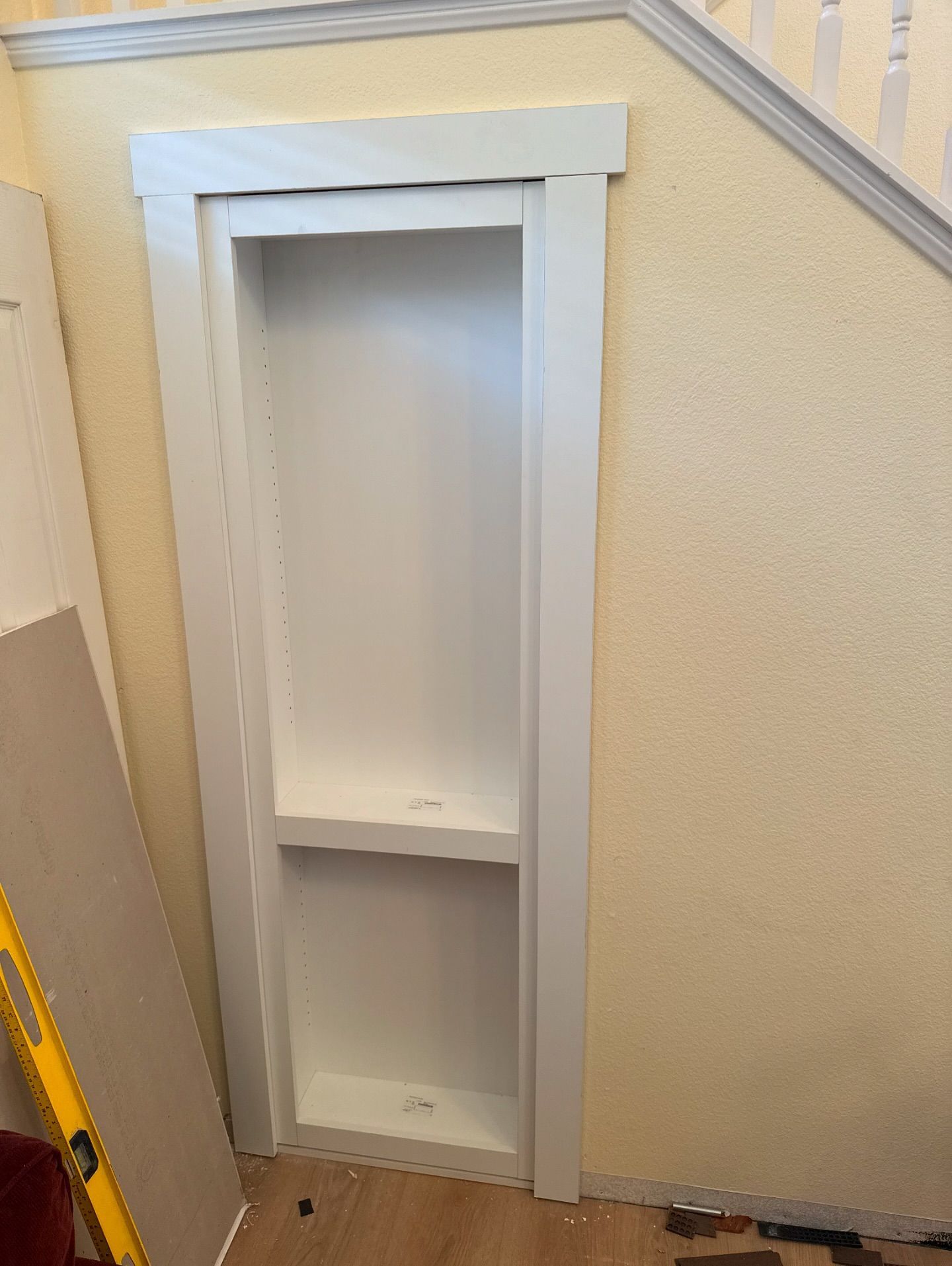 White built-in wall shelf unit with two shelves, framed with white molding, set into a yellow wall, near a staircase.