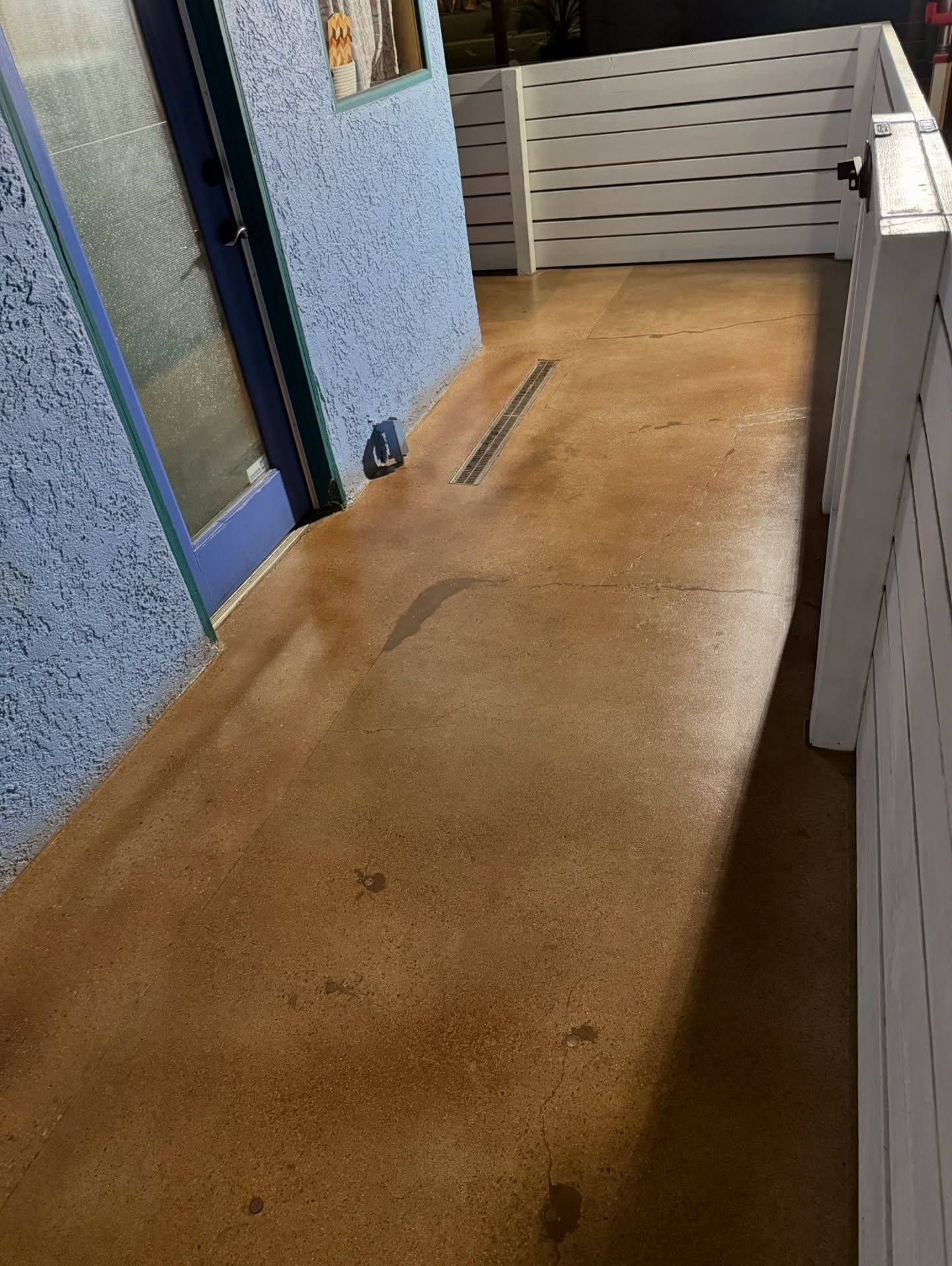 Brown concrete walkway with blue stucco wall, white slatted fence, and a door.