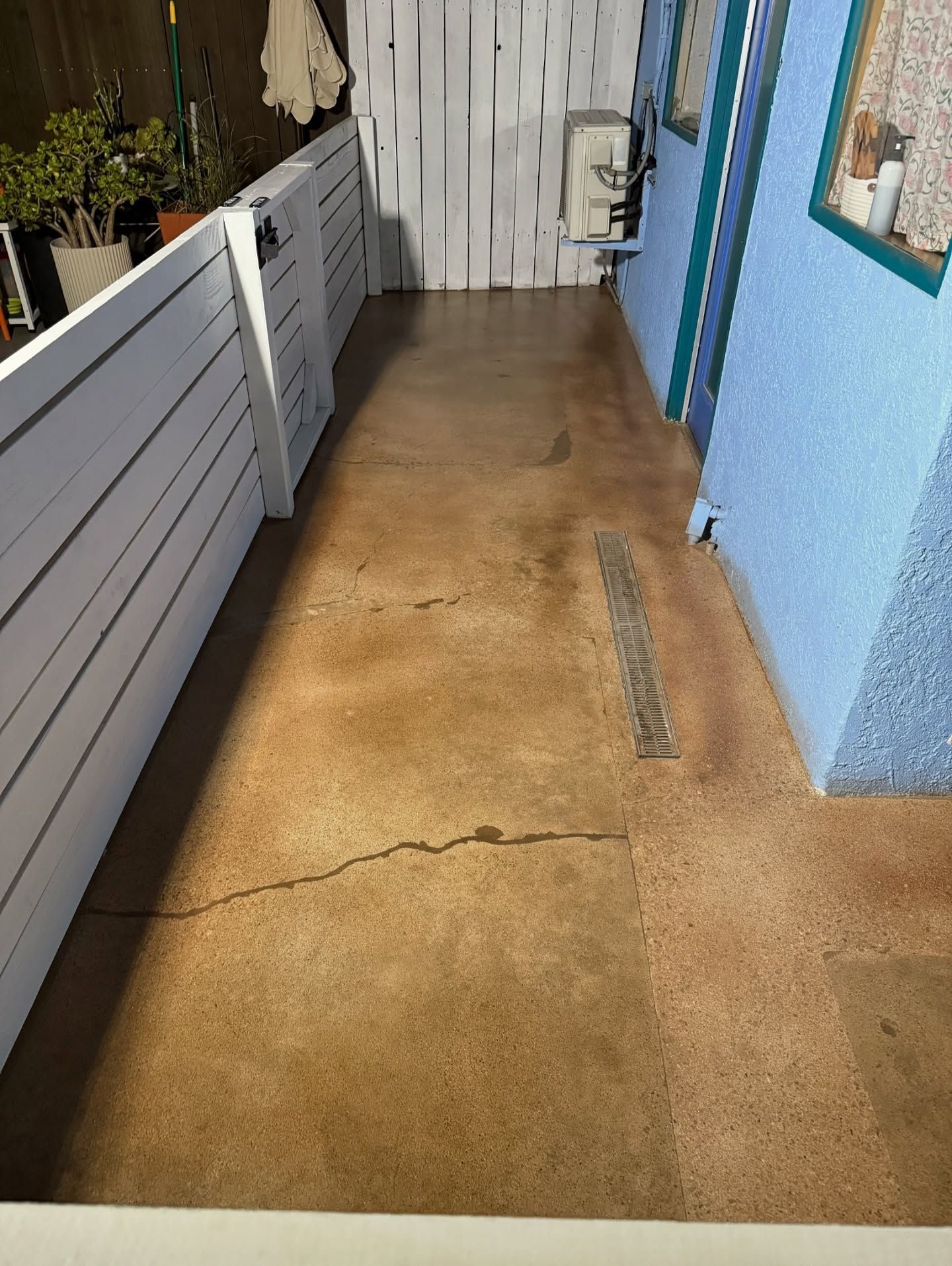 Concrete patio with brown staining and cracks, bordered by white fence and blue stucco wall.