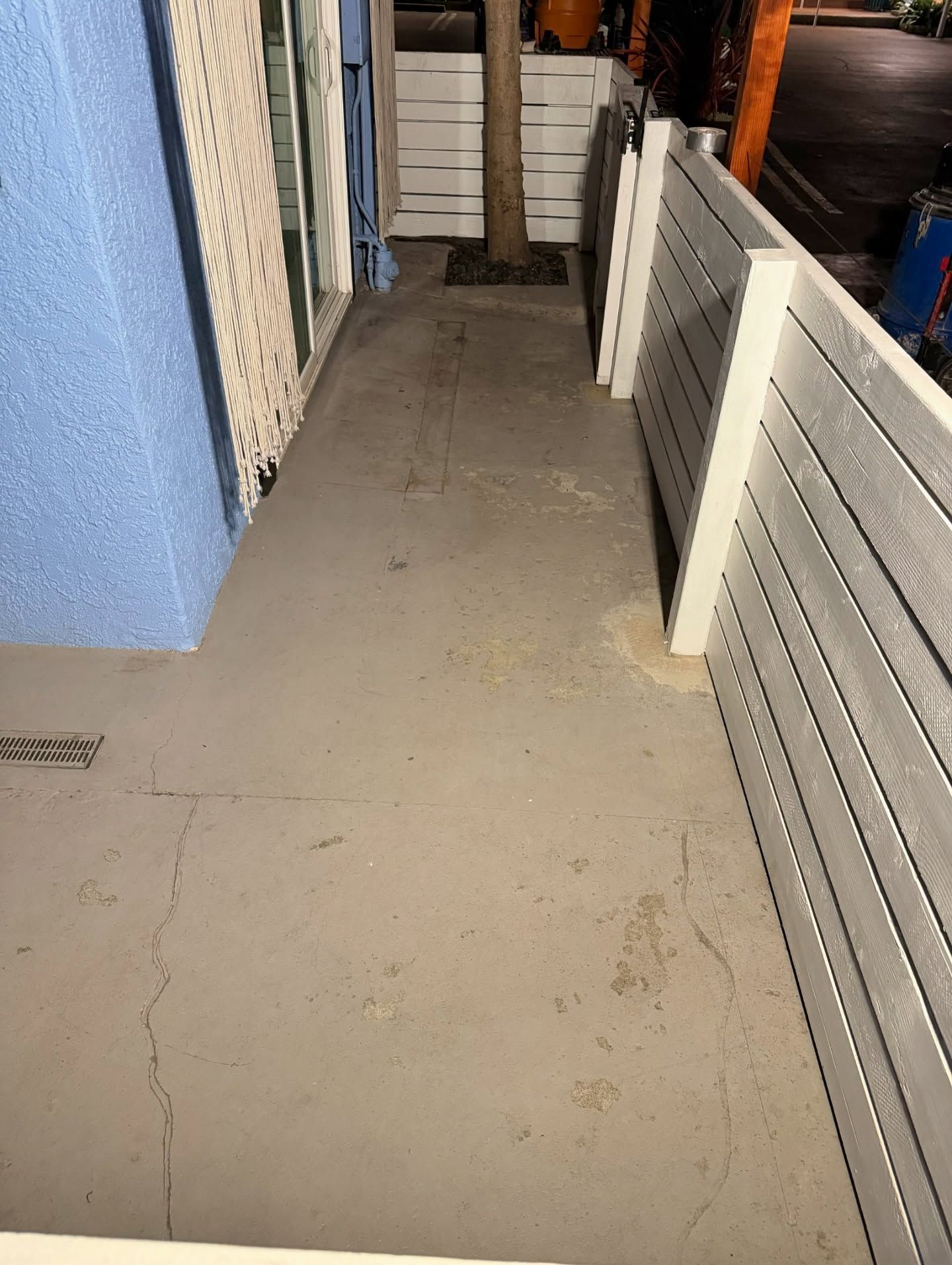 Concrete walkway between a blue wall and white slat fence, with a tree in the background.