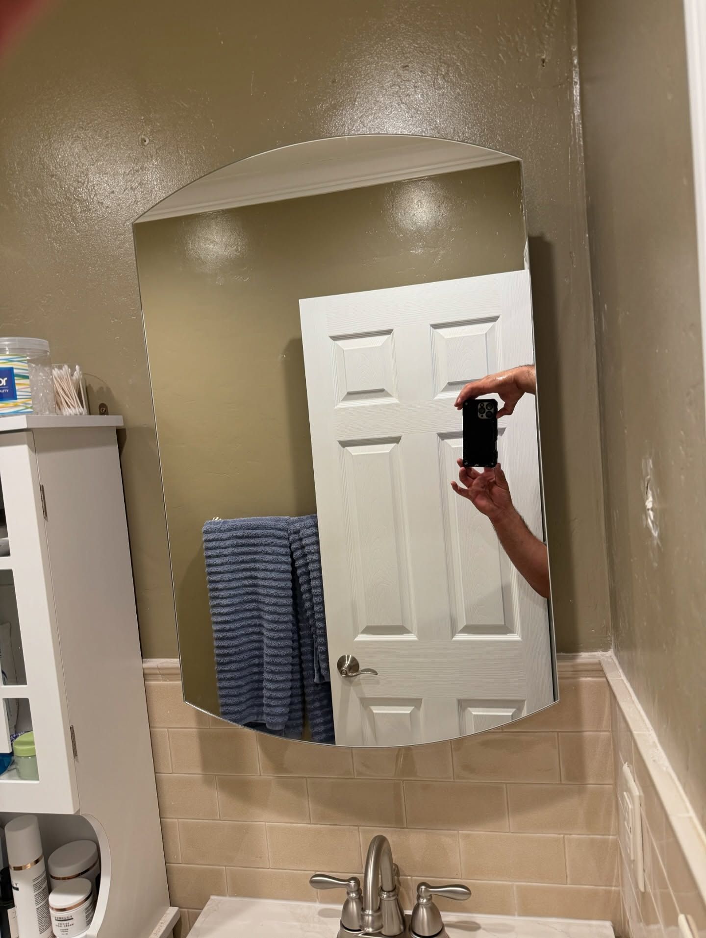Mirror in a bathroom reflects a closed white door and a person holding a phone. A towel hangs nearby.