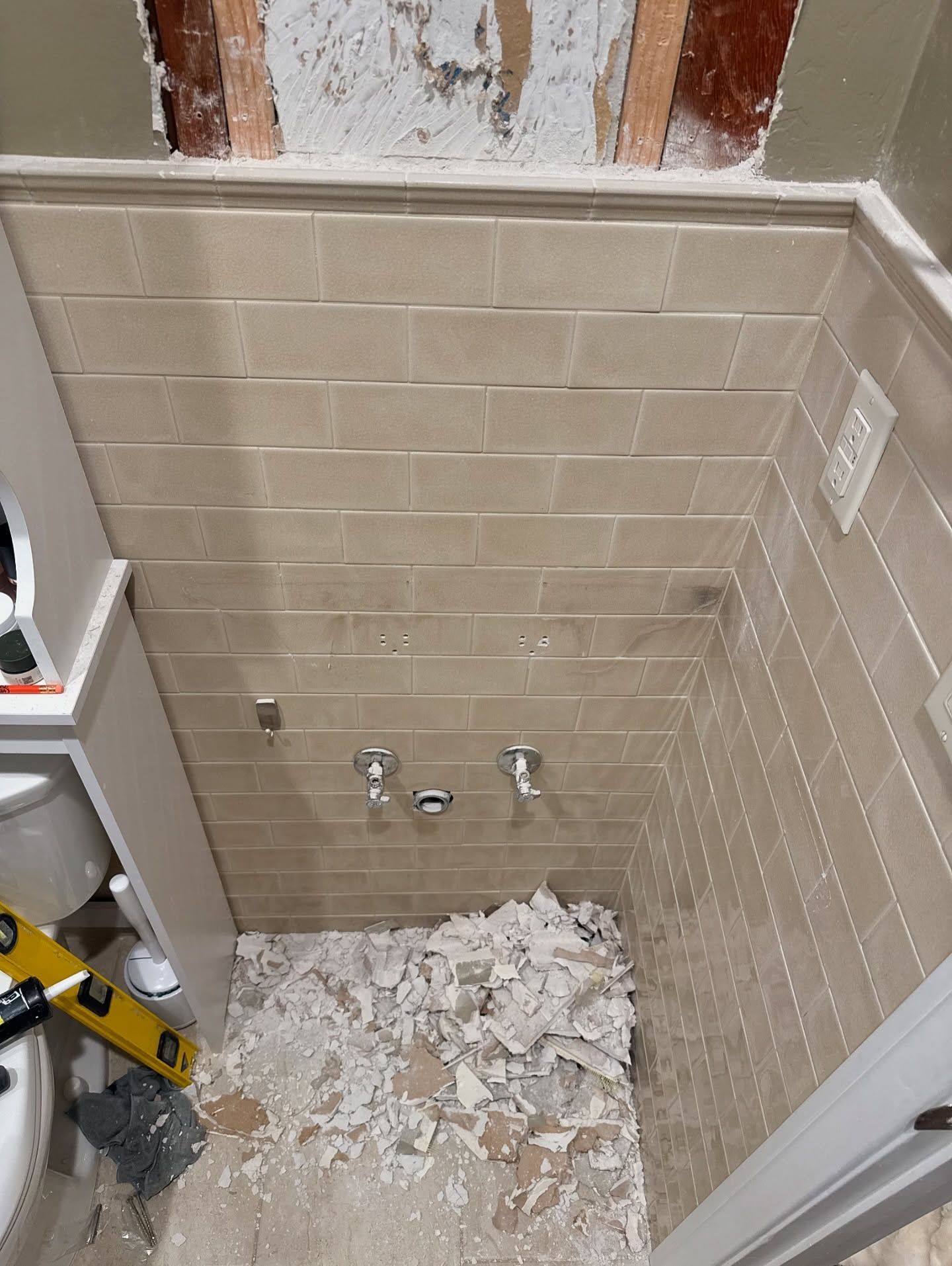 Bathroom wall with beige tiles, plumbing, and debris on the floor, under construction.