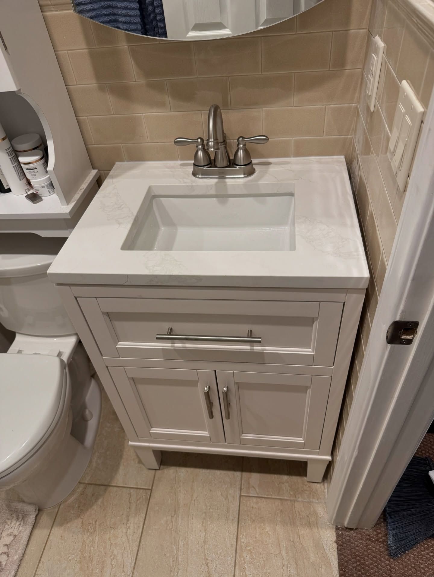 White bathroom vanity with sink, faucet, and cabinet doors. Beige tile and a round mirror in the background.
