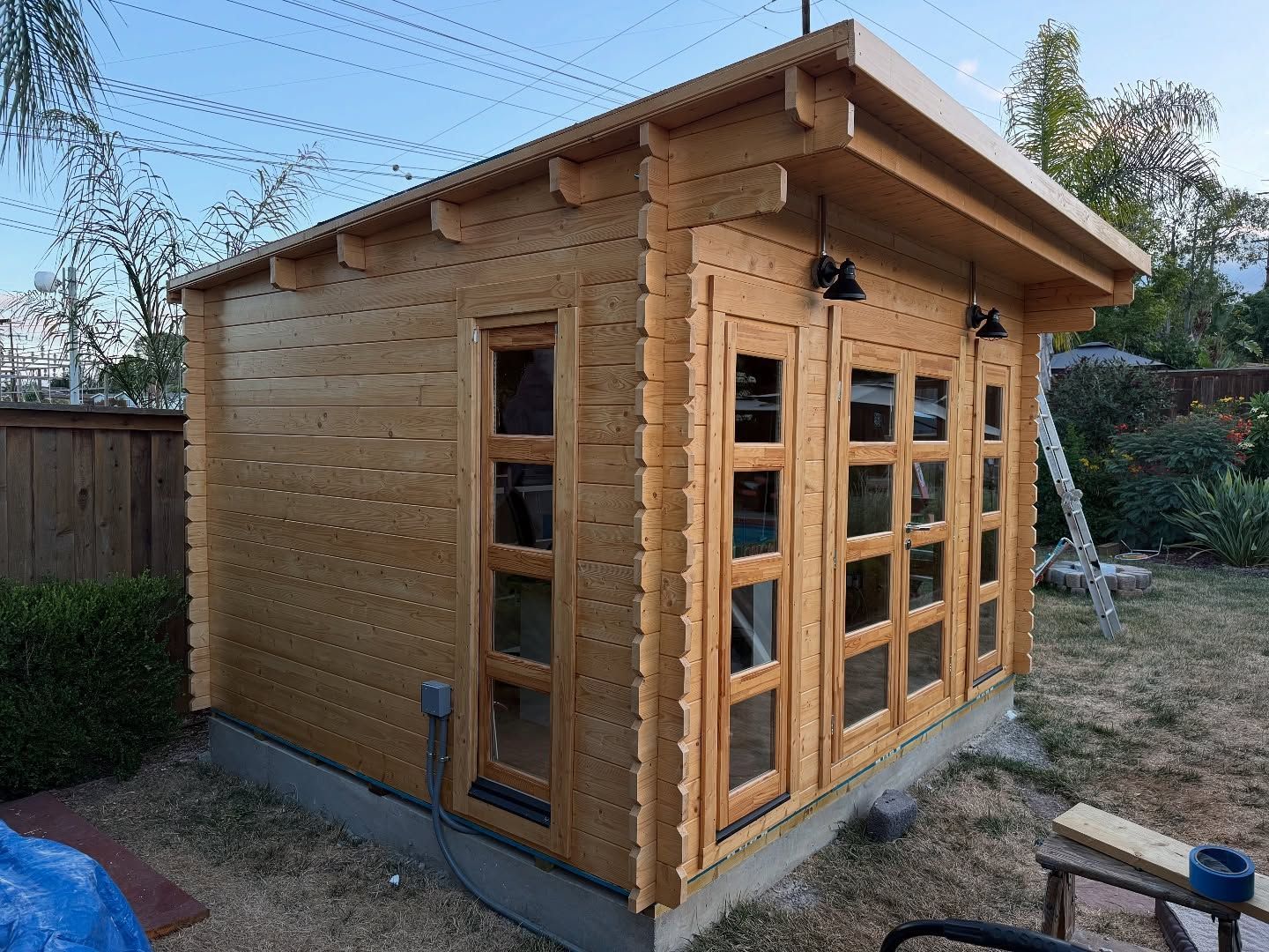 Wooden backyard shed with windows and doors, surrounded by grass and a fence.
