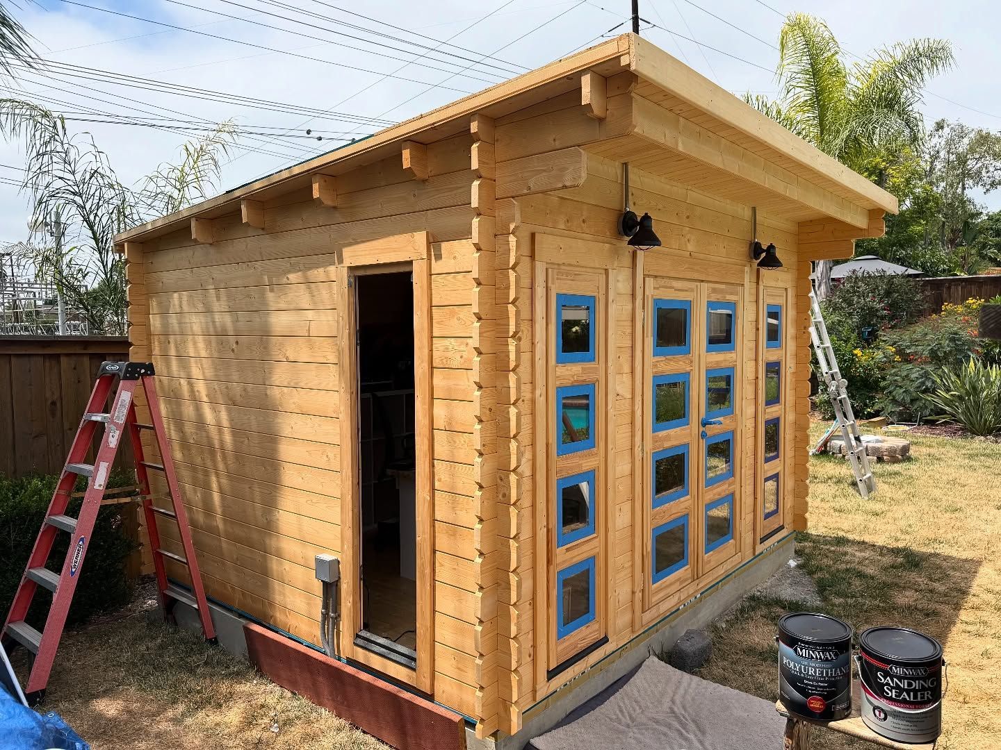 Wooden shed under construction; door and windows visible; blue trim; ladder nearby.