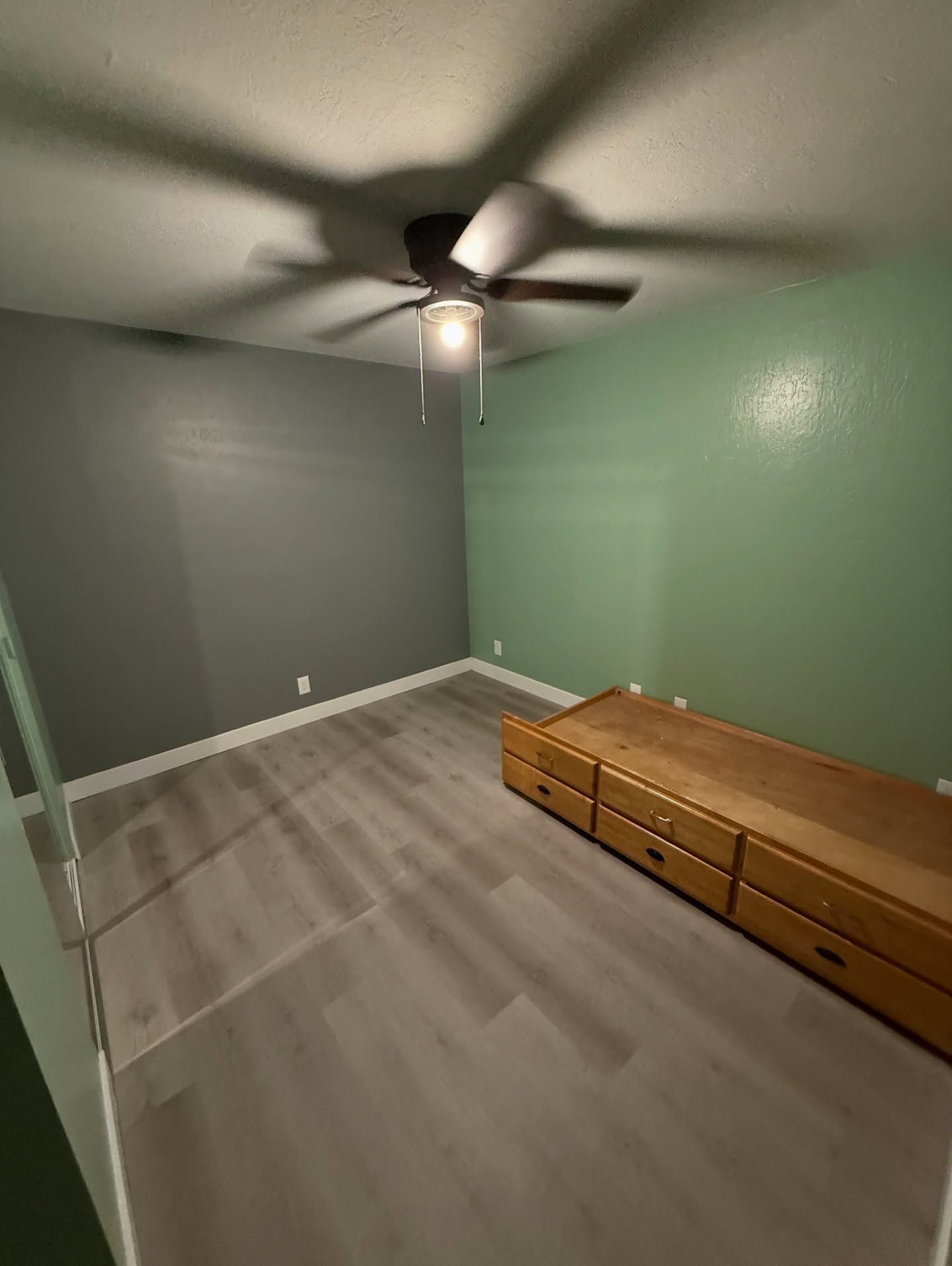 Empty bedroom with gray and green walls, wood dresser, light gray floor, and ceiling fan.