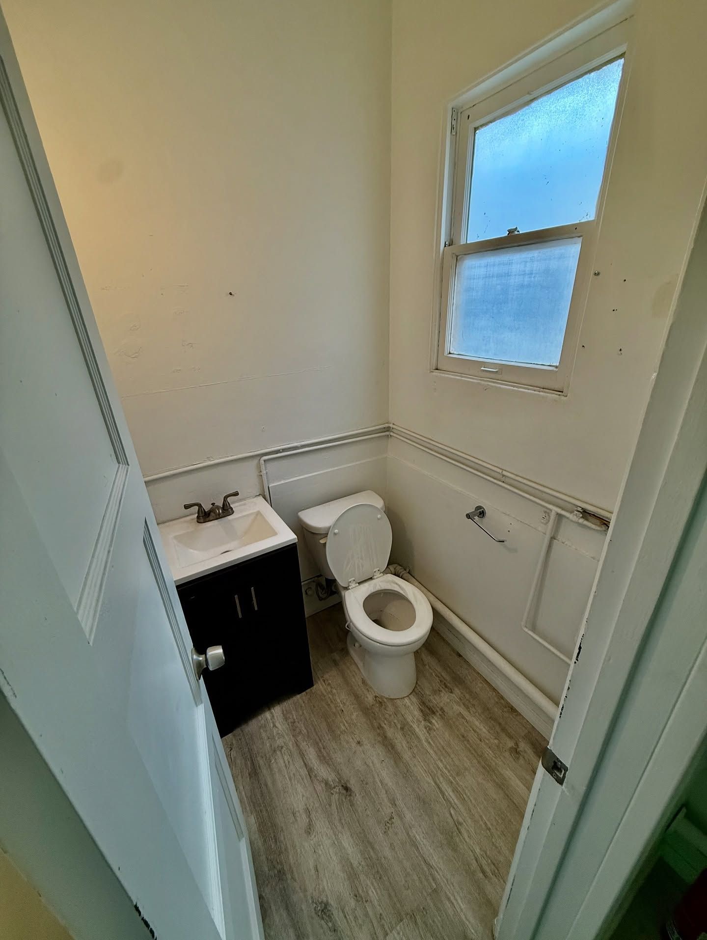 Small bathroom with a toilet, sink, and window. White walls, wood-look floor, black cabinet.