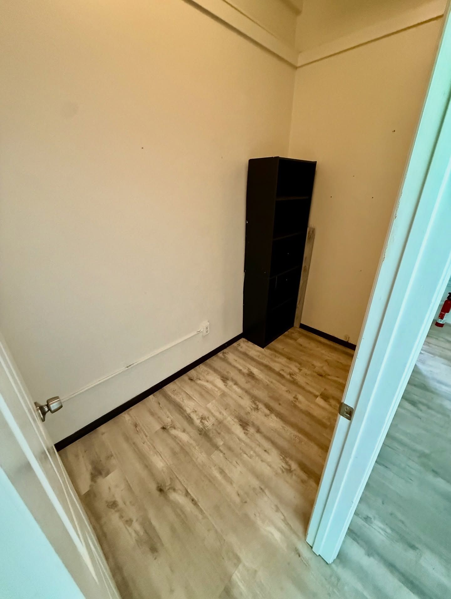 Small empty room with light wood-look flooring and a black bookcase in the corner.