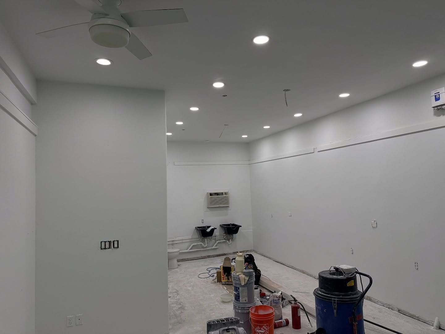 White-walled room with recessed lighting, ceiling fan, two sinks, and construction materials on the floor.