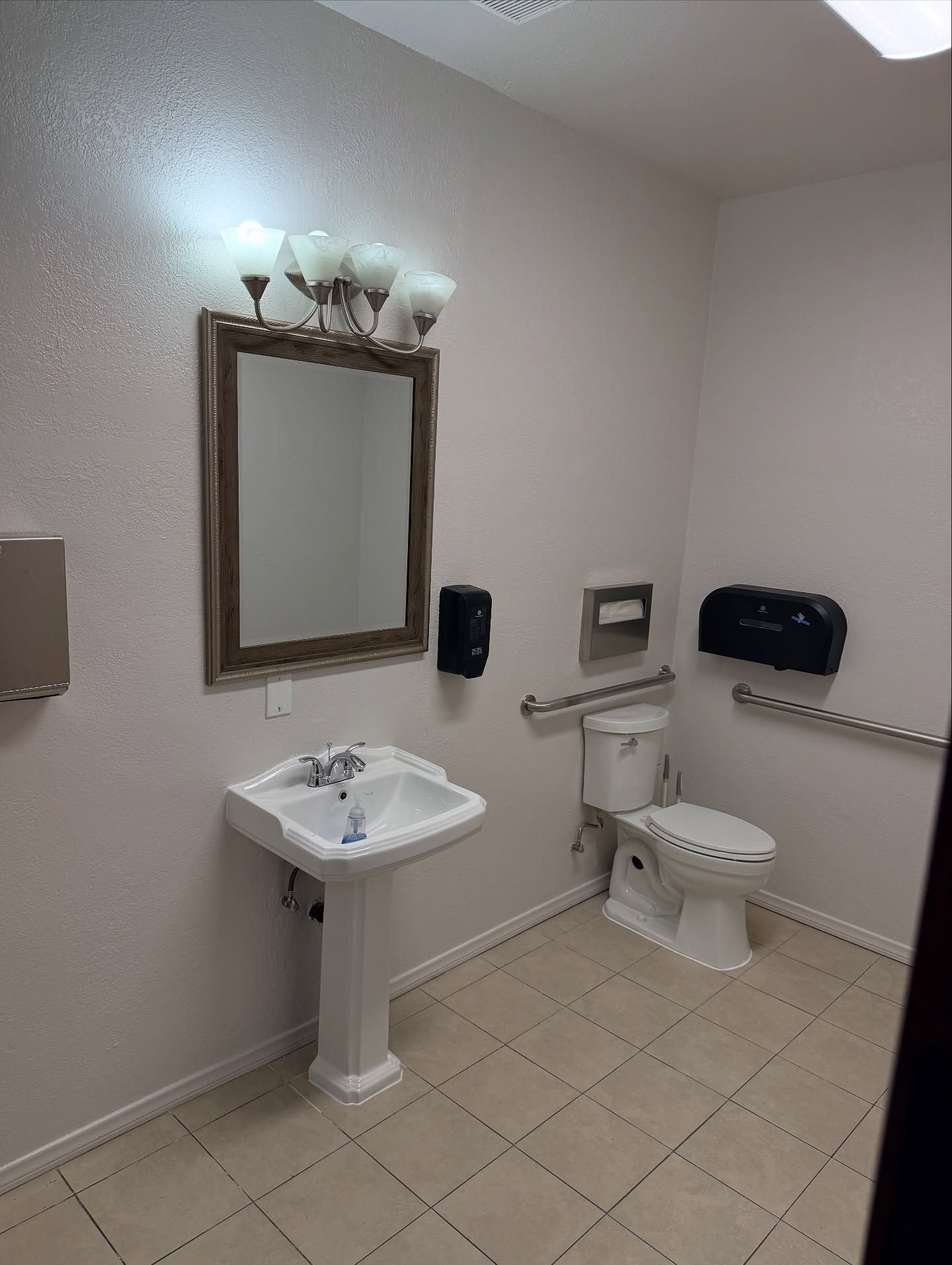 Bathroom with sink, mirror, toilet, and grab bar. Light walls, tile floor, and paper towel dispenser.