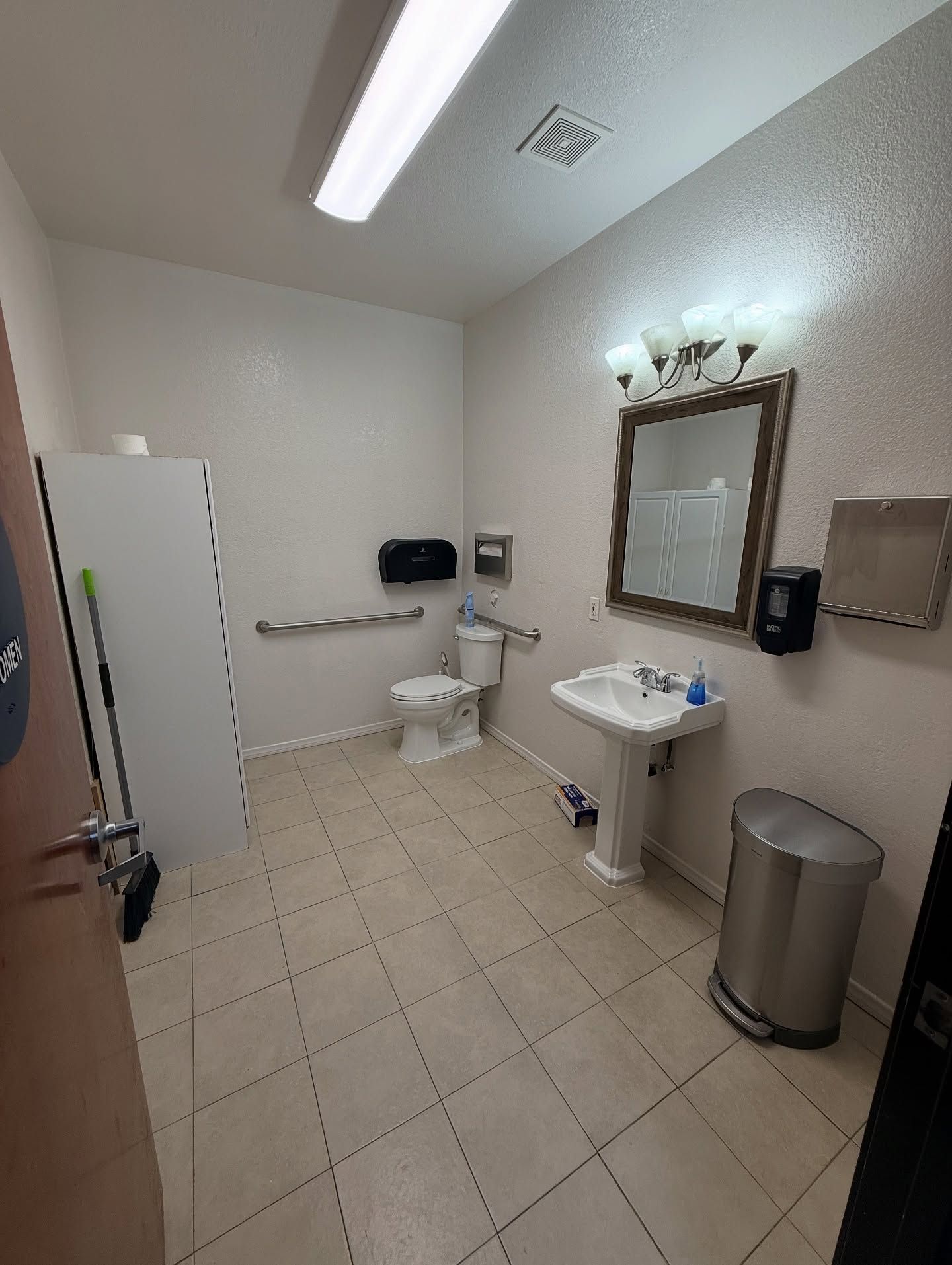 Small accessible restroom with a toilet, sink, mirror, and trash can. Features a grab bar.