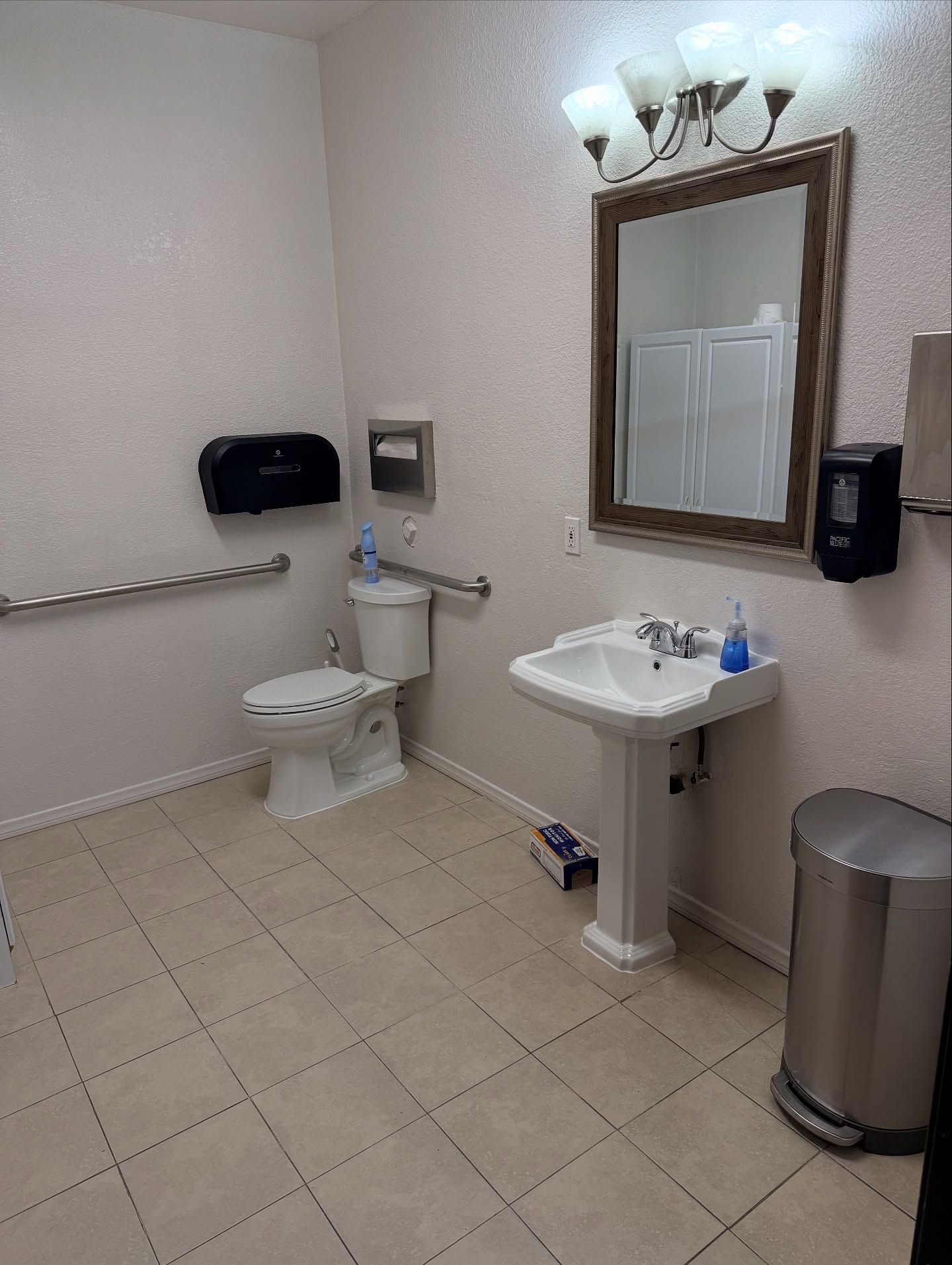 ADA-accessible public restroom with a toilet, sink, mirror, and grab bars. Beige tile floor, light walls.