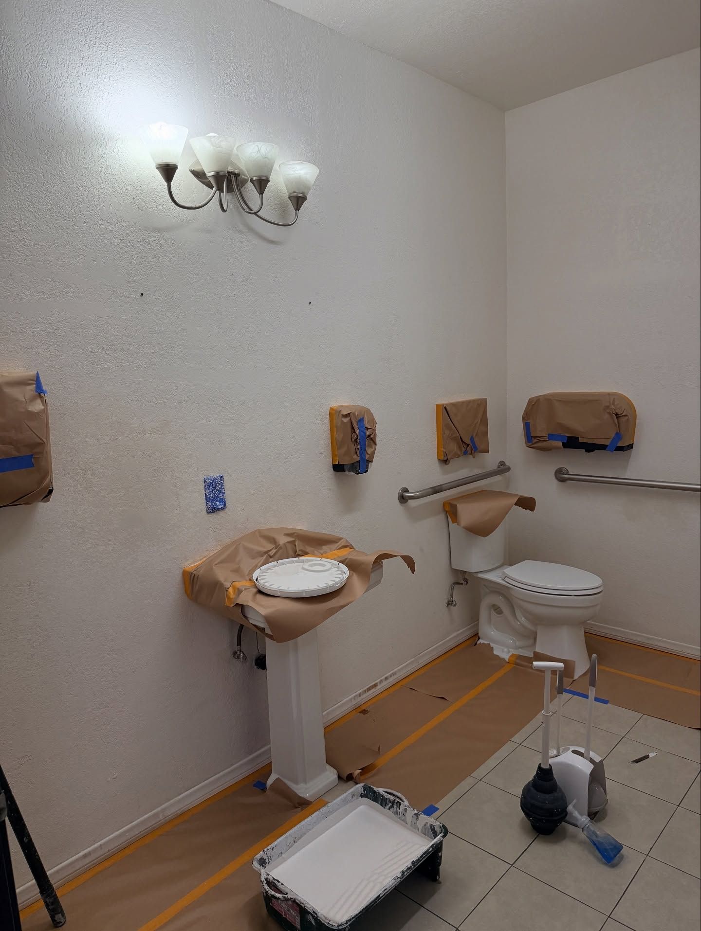 Bathroom being painted, fixtures covered with paper, paint roller and tray on the floor.