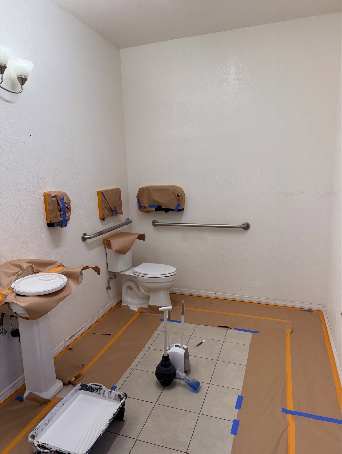 Bathroom being painted, with toilet, sink, and safety bars. Walls are white, floor has protective covering.