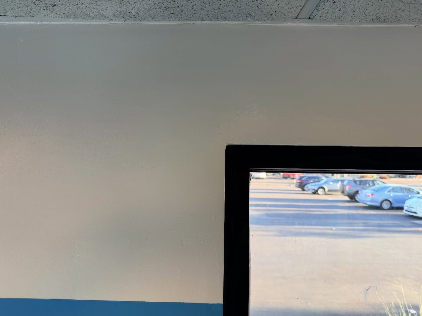 View of a parking lot through a black-framed window. White wall and blue stripe below.