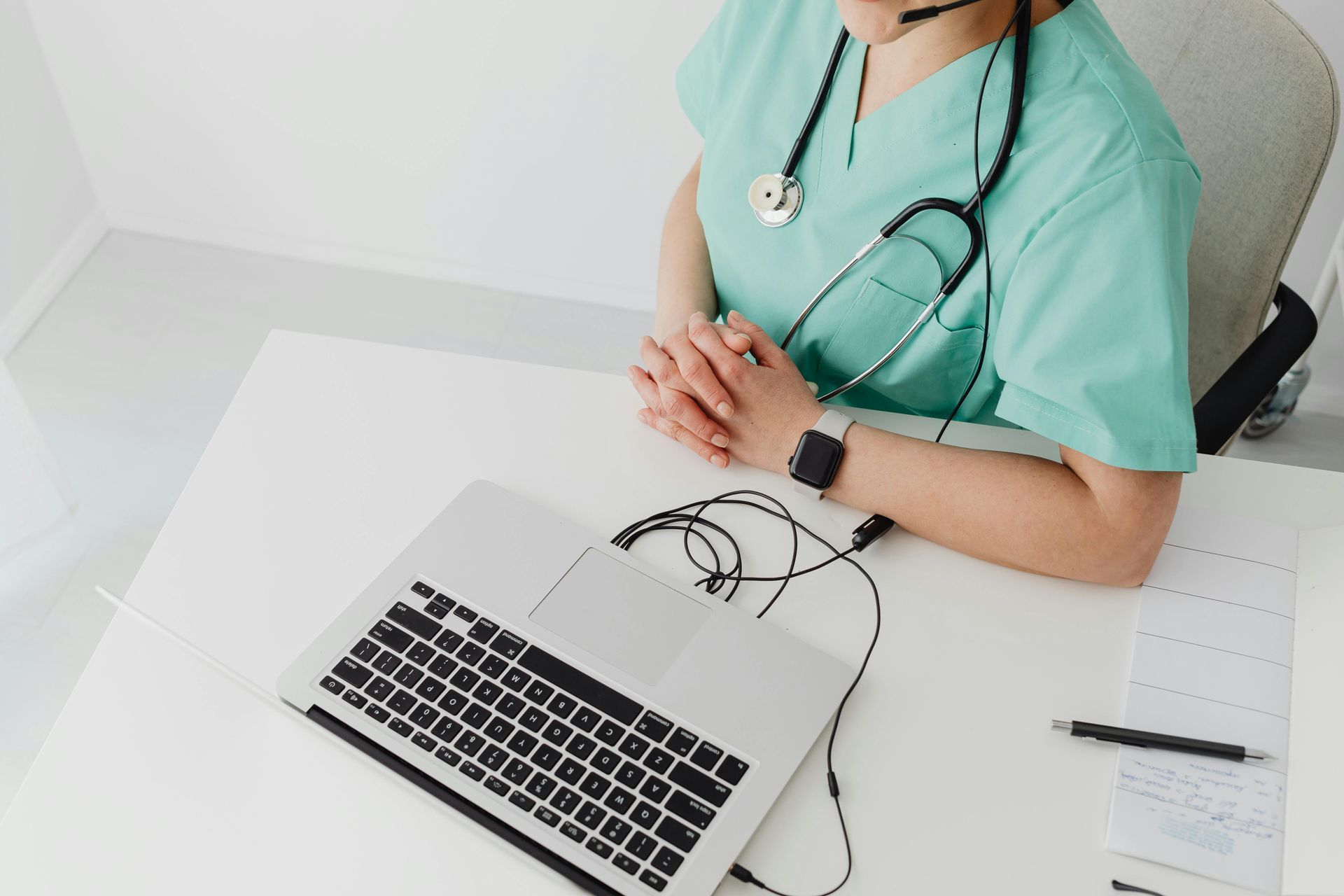 A doctor is sitting at a desk with a laptop and a stethoscope around her neck.