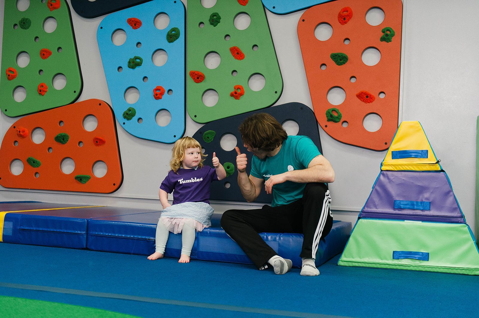 Children in a gymnasium gather around a green mat, with a teacher, interacting and observing.