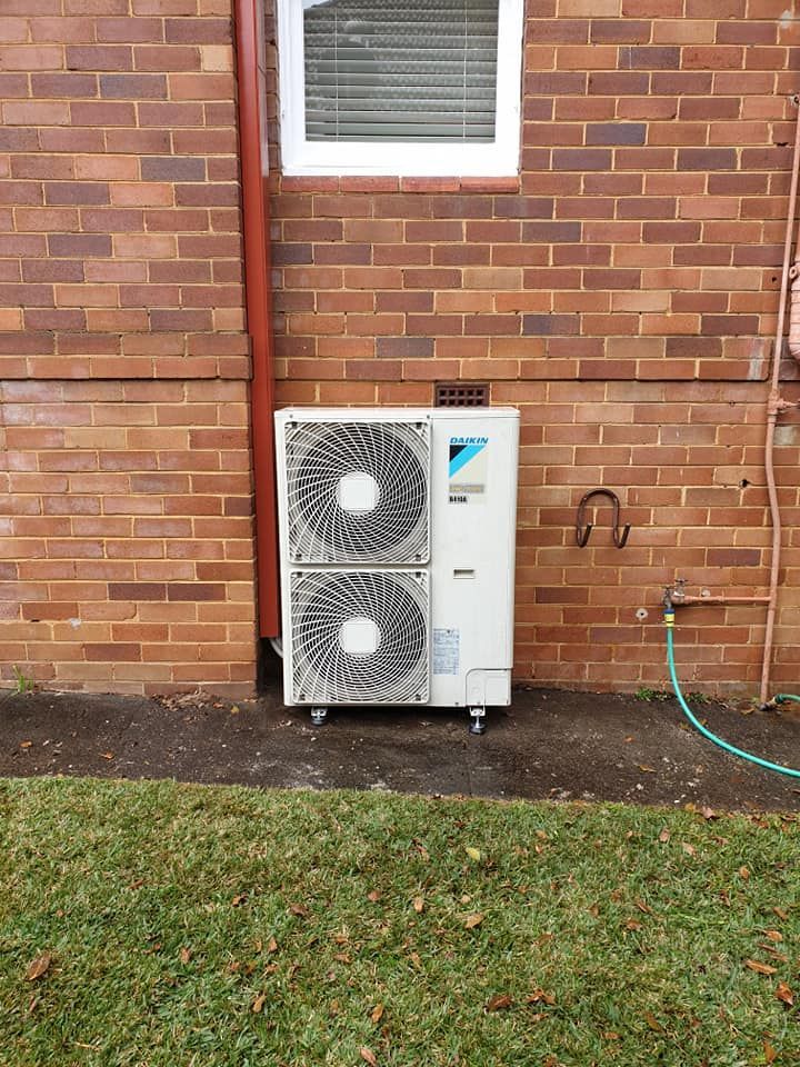 An air conditioning unit mounted on a brick wall, below a window, beside a grass lawn.
