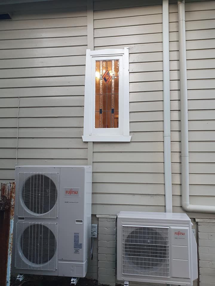 Two air conditioning units mounted below a small window on a light-colored house wall with white trim.
