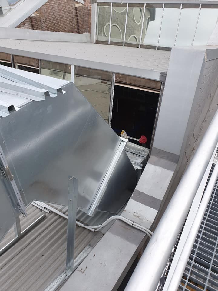 Metal air duct on building rooftop, light gray, angled toward an opening.