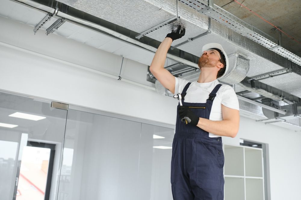 Construction worker in overalls and hard hat inspecting overhead ductwork in a building.