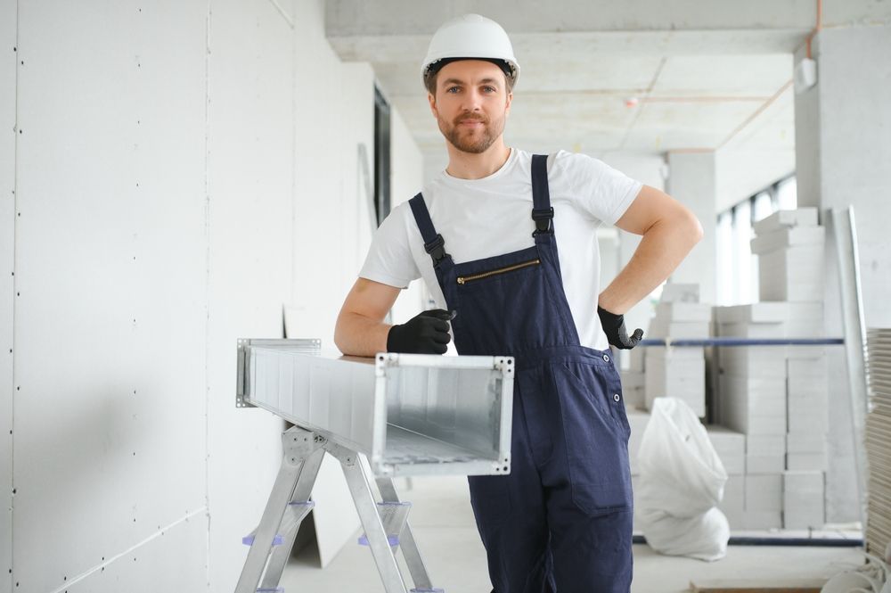 Construction worker in a white hard hat and overalls, leans on a ventilation duct. Interior construction site.