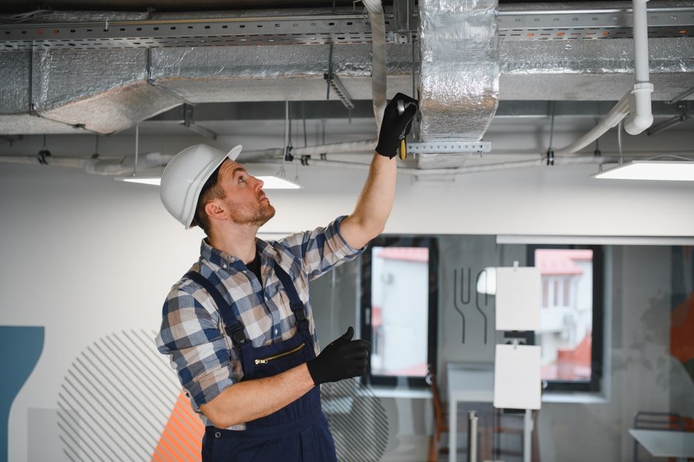 Man in hard hat and work clothes inspecting ductwork in a building.