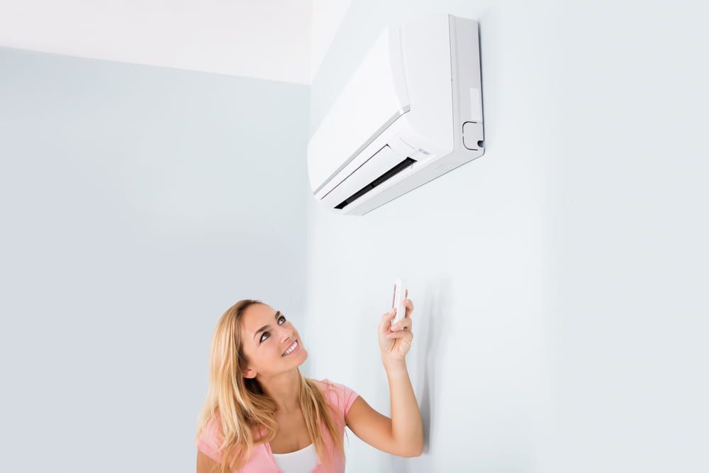 Woman smiling, using remote to adjust a white air conditioner on a light blue wall.