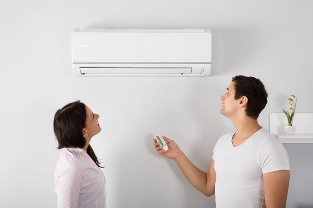 Man adjusts AC unit with remote as woman watches, both looking up in a white room.