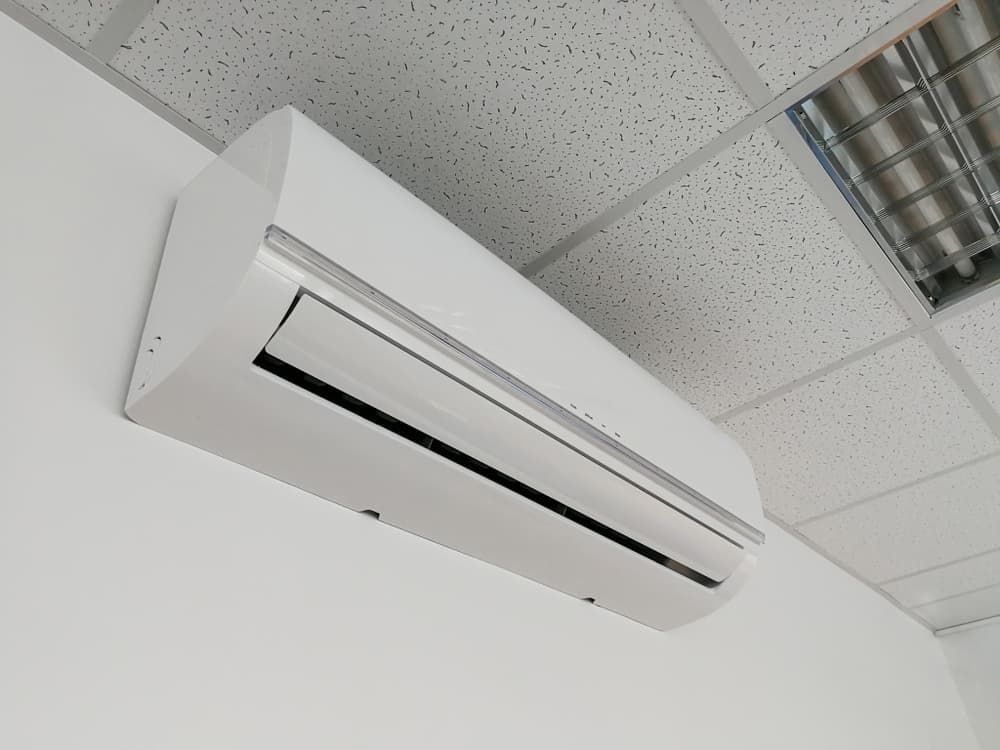 White wall-mounted air conditioner on a white wall, beneath a drop ceiling with recessed lighting.