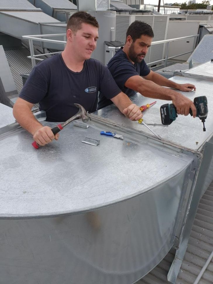 Two men working on a metal structure, one hammering and the other using a power drill on a rooftop.