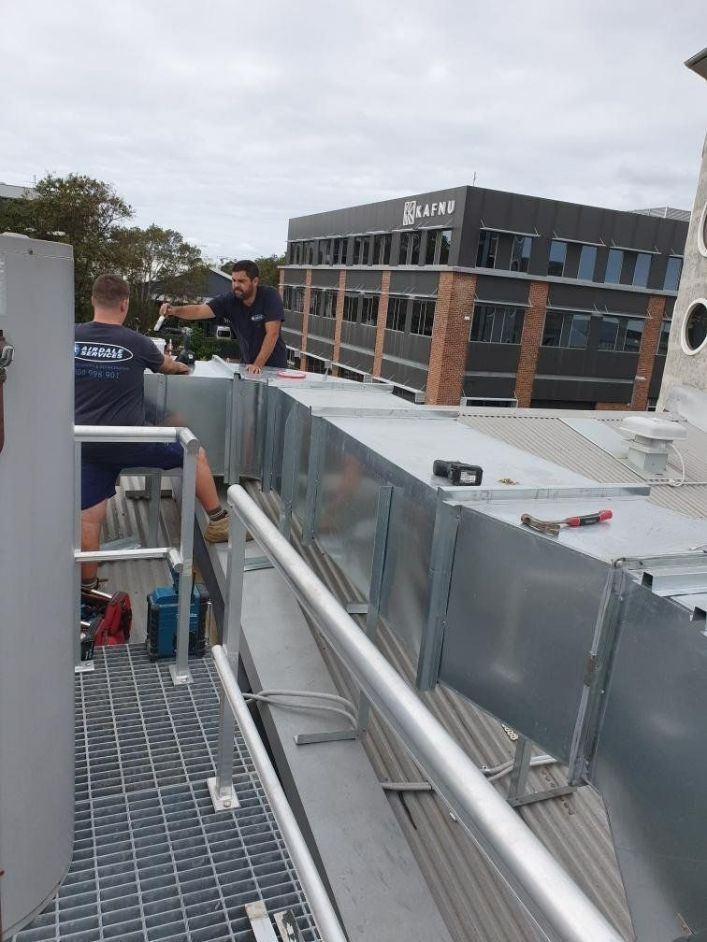 Two workers installing metal ductwork on a rooftop, with an office building in the background.