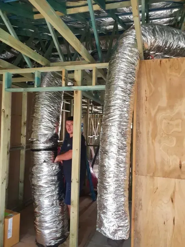 Person standing inside wooden frame construction, surrounded by flexible silver air ducts.