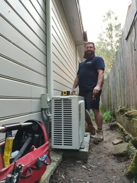 Man in uniform stands next to an air conditioner, toolbox nearby, between a building and a fence.