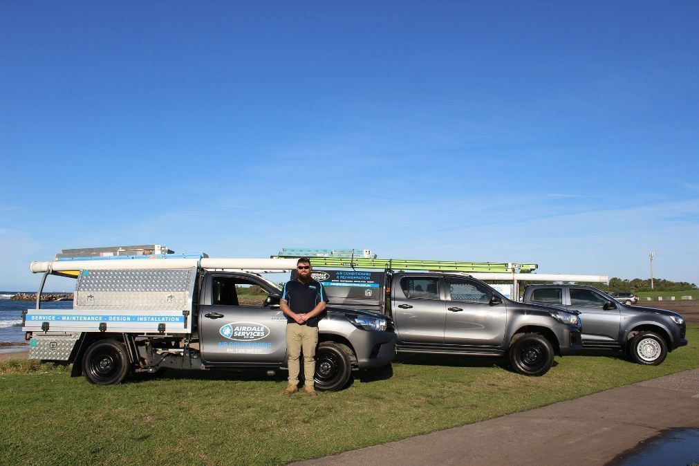 Man stands in front of three trucks parked on grass near water under a blue sky.