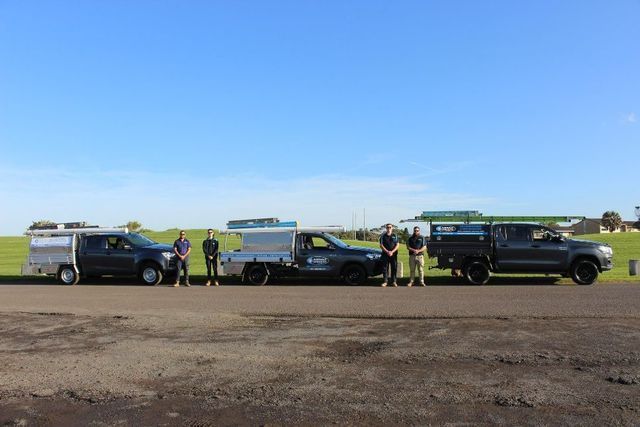 Four trucks with workers standing in front, on a paved area, with green field and blue sky background.