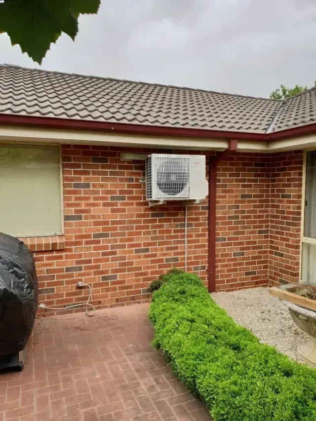 An air conditioner unit mounted on a red brick wall of a house next to a green hedge and a tiled patio.