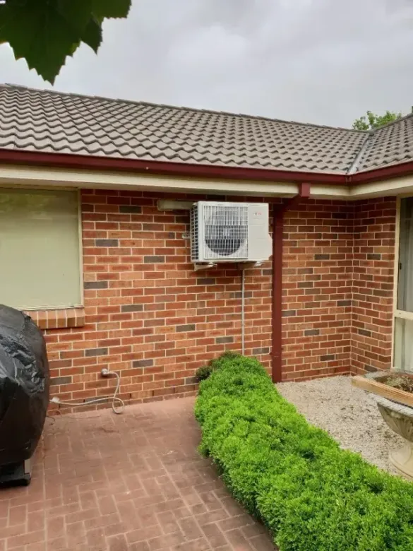 Air conditioning unit mounted on a red brick exterior wall under a tiled roof. A green hedge is below.