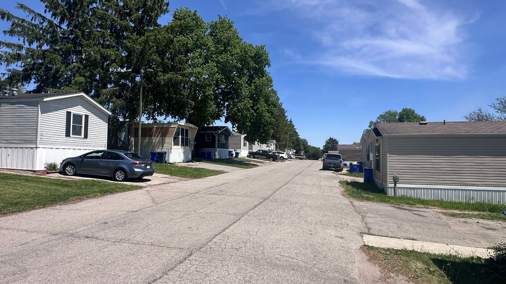 Quiet residential street with mobile homes, parked cars, and trees under a blue sky