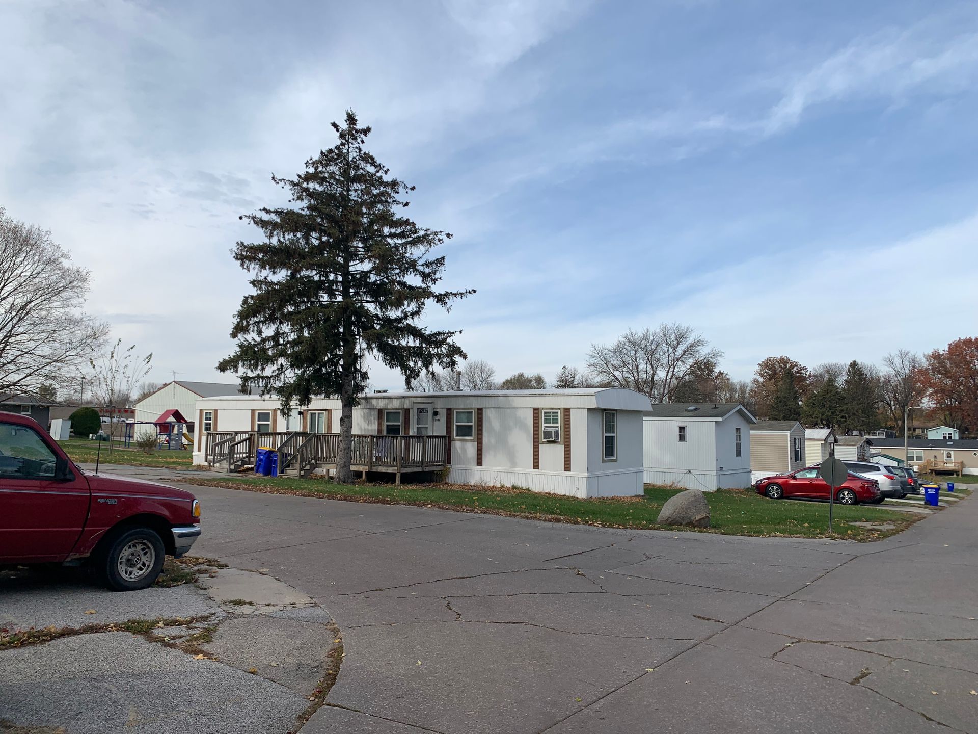 Mobile home park with a large pine tree, parked cars, and cloudy sky