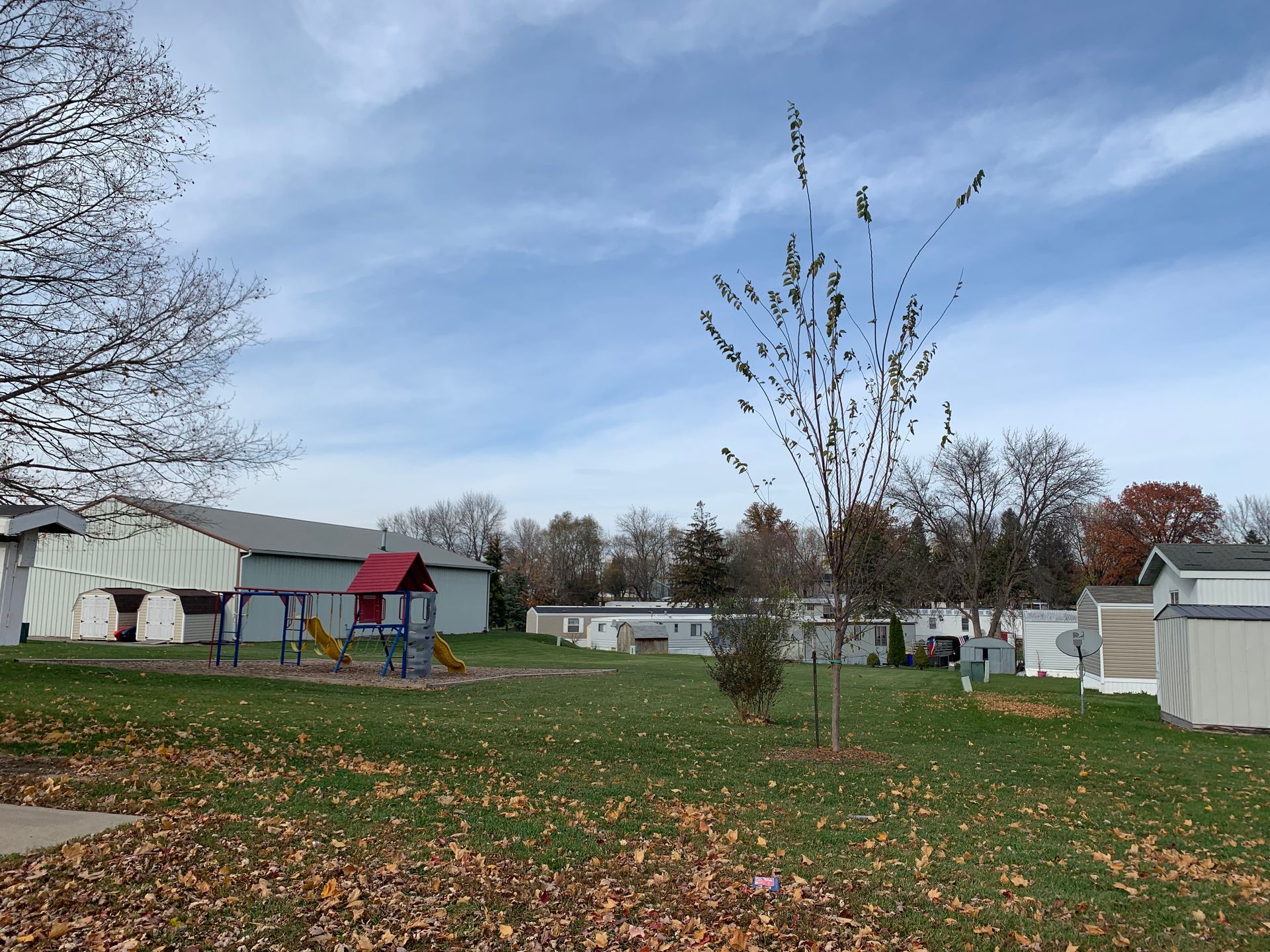 Mobile home park with grassy lawn, young trees, and a small playground under a cloudy sky