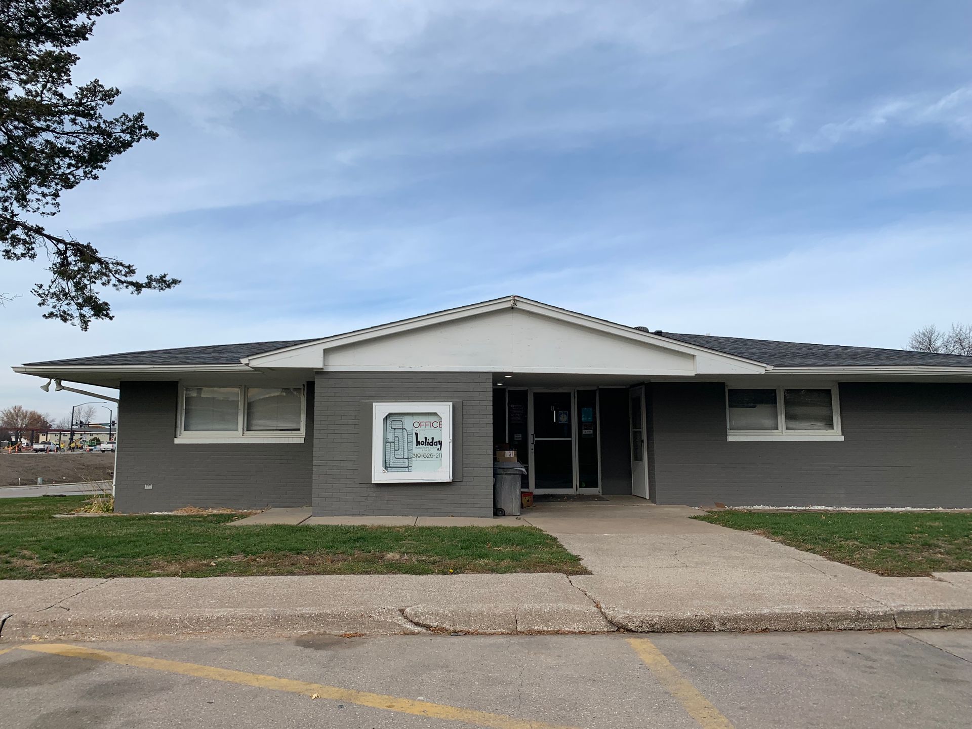 Single-story gray building with a central entrance, small windows, and a concrete walkway under a cloudy sky